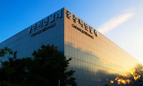 蓝天下的现代玻璃建筑外墙,顶部可见韩文及 GWANGJU BIENNALE 字样。