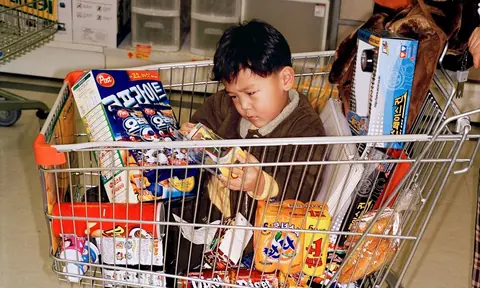 一名儿童坐在装满彩色包装商品的购物车中翻看物品。
