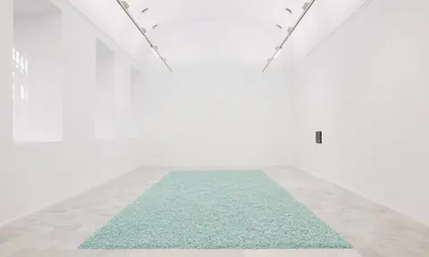 纯白展厅地面中央铺着一块浅青色颗粒状长方形装置。