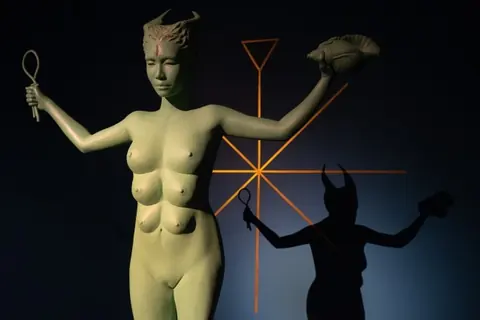暗色展厅中一尊多乳、双臂持物的人形雕塑及其墙面投影
