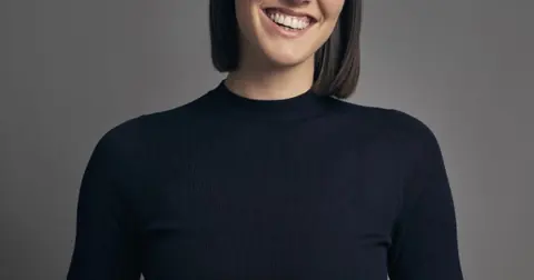 灰色背景前一名短发女性的半身肖像,人物身穿黑色上衣并面带笑容。